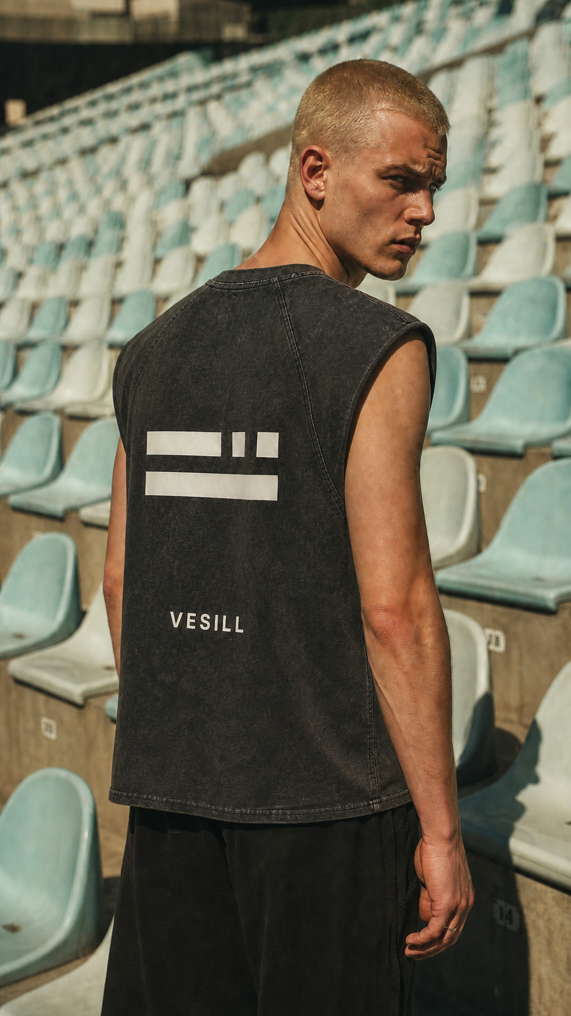 Person wearing a black sleeveless shirt with a logo and text in an outdoor setting with stadium seats.